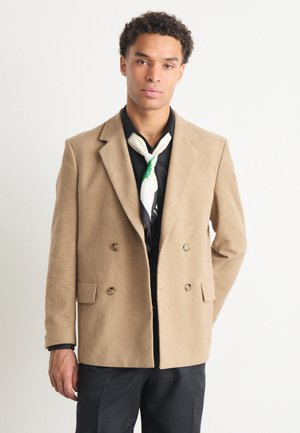 Beige double-breasted blazer made of textured fabric, featuring two front pockets and gold-tone buttons, paired with a black shirt and green-accented scarf.