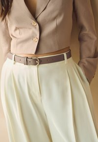 Ceinture en cuir marron avec une boucle en métal, portée par-dessus un pantalon plissé jaune pâle, associée à un blazer court beige.