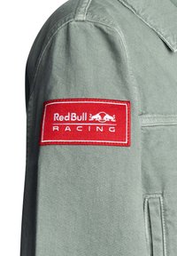 Parche con el logo de Red Bull Racing en blanco sobre un fondo rectangular rojo, cosido en la manga de tela gris claro.