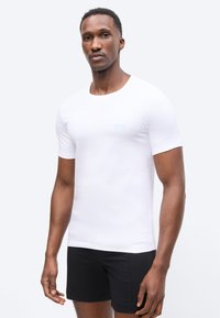 Mand iført tætsiddende hvid skjorte med korte ærmer og sorte shorts, stående foran en ensartet lys baggrund, vendt fremad.