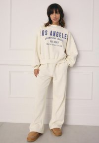Cream-colored oversized sweatshirt with blue text, paired with matching wide-leg pants and tan slip-on shoes. Smooth fabric and relaxed silhouette.