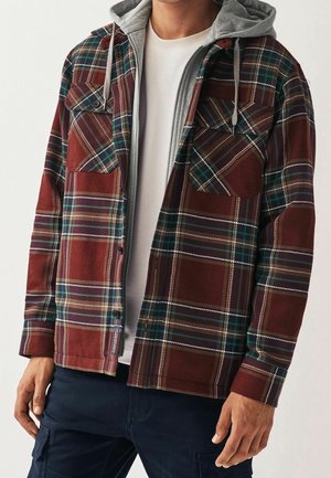 Mann trägt eine rot-grün karierte Jacke mit Knöpfen über einem grauen Zip-up-Hoodie und einem weißen Shirt, kombiniert mit dunkler Hose.