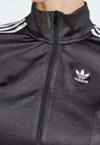 Svart zip-up jacka gjord av slät material, med vitt Adidas-logotyp och tre vita axelränder. Hög krage och texturerad tyg.