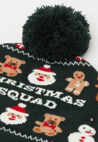 Zielona, dziergana czapka z miękkim pomponem, ozdobiona świątecznymi wzorami z niedźwiedziami, Mikołajem i piernikowymi ludzikami w kontrastujących kolorach i wzorach.