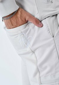 Lichtgrijze broek met een gestructureerde afwerking, voorzien van een zijzak en een wit geborduurd logo. Glad stof en subtiele ontwerpelementen.