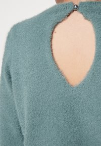 Suéter de terciopelo verde azulado con diseño de ojal en la espalda y cierre de botón sencillo. Textura suave con un corte holgado que resalta el escote.