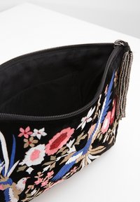Czarna torebka kopertowa z kolorowym haftem w kwiaty i ptaki, otwarty zamek błyskawiczny oraz srebrny chwost z koralików na białej powierzchni.