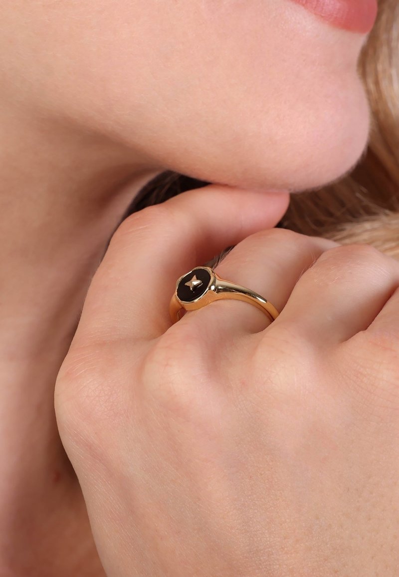 Gros plan d'une main reposant près du menton, portant une bague en or avec une pierre précieuse noire et un petit détail en relief au centre.