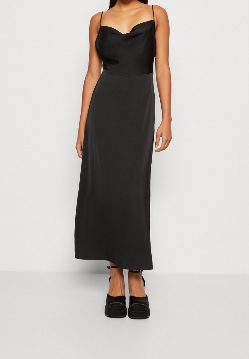 Femme portant une robe nuisette noire en satin sans manches avec un col bénitier et des talons plateformes noirs, debout devant un fond clair uni.