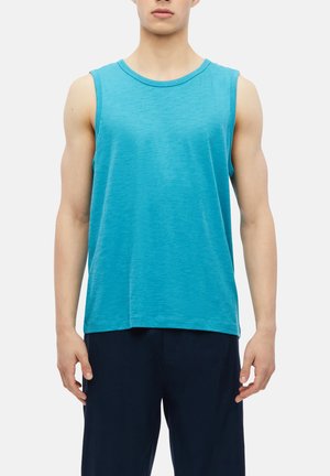 Homme portant une chemise sans manches bleu clair et un pantalon bleu foncé, debout, les bras relâchés le long du corps devant un fond uni.