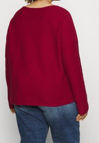 Pull en tricot rouge avec une coupe décontractée, un large col et des épaules tombantes. Associé à un jean en denim bleu, mettant en avant un style décontracté.
