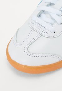 White leather sneaker close-up with orange rubber outsole, white laces, and contrasting stitching details, featuring a streamlined shape.