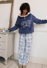 Blå fleece pullover med hvid krave og 'APRÈS' grafik, parret med lyse ternede pyjamasbukser og beige slippers, placeret i et lyst rum.