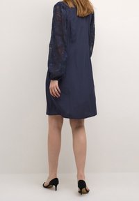 Marineblå klänning med genomskinliga spetslånga ärmar, avslappnad passform, mjuk textur, knälång och med subtil lyster, kombinerad med svarta högklackade skor.