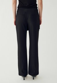 Melni augstās vidukļa bikses ar taisno kāju dizainu, kurām ir divas aizmugures kabatas un gluda tekstūra. Valkātas ar melniem papēžiem.