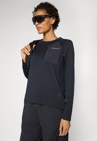 Maglietta sportiva a maniche lunghe di colore blu scuro con una tasca sul petto nera a contrasto e logo. Texture liscia con una vestibilità comoda, abbinata a pantaloni scuri.