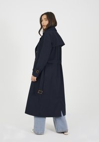 Granatowy trencz z szerokim kołnierzem, zapinany na guziki z paskiem w talii. Posiada pasujący materiałowy pasek i mankiety z kwadratowymi akcentami z metalu.