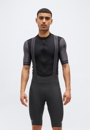 Muž v černém a šedém krátkém cyklistickém oblečení s nohavicemi na zádech, stojící před jednoduchým světle šedým pozadím.