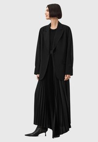 Blazer nero su misura sopra una gonna maxi plissettata nera, con pieghe verticali, abbinato a stivali alla caviglia a punta. Tessuto dalla texture liscia.