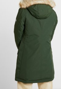 Grüne Parka mit fellgefütterter Kapuze, langer Passform und Seitentaschen; glatte Textur, minimale Nähte und leicht taillierter Schnitt.