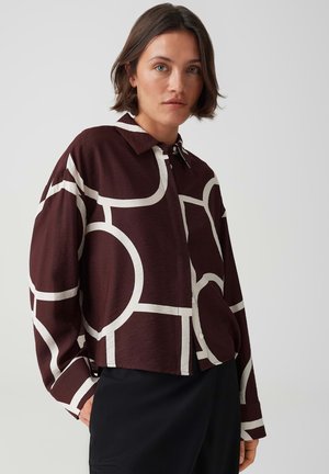 Frau mit kurzen braunen Haaren trägt ein dunkelweinrotes Hemd mit großen weißen geometrischen Mustern und schwarze Hosen.