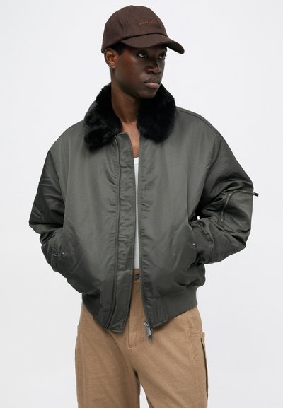 Zaļš bombdera stils jaka ar melnu mākslīgās kažokādas apkakli, rāvējslēdzēja aizdari un sānu kabatām. Valkāta virs baltas blūzes un kombinēta ar brūnām biksēm.