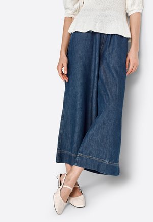 Mujer con vaqueros de pierna ancha y corte cropped, y una blusa de punto color crema, de pie con las piernas cruzadas, usando zapatos planos blancos con tiras.