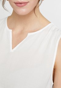 Blouse blanche sans manches avec un décolleté en V, un tissu léger et de délicates fronces aux épaules pour une coupe décontractée.