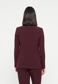 Bordeaux blazer met een getailleerde pasvorm, voorzien van lange mouwen en een klein embleem op de achterkant. Glad textuur en gestructureerd ontwerp.