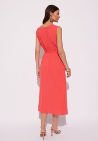 Koronkowa sukienka midi w kolorze koralowym z dopasowaną talią, bez rękawów i subtelnymi pliskami z tyłu. W zestawie z cielistymi szpilkami i dużymi kolczykami.