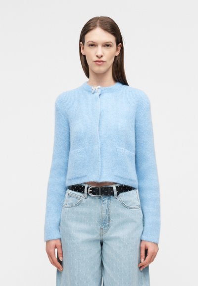 Γυναίκα που φοράει ανοιχτό μπλε χνουδωτό ζακέτα με μπροστινές τσέπες, συνδυασμένη με ανοιχτόχρωμο ψηλόμεσο τζιν και μαύρη ζώνη με τρουκς.