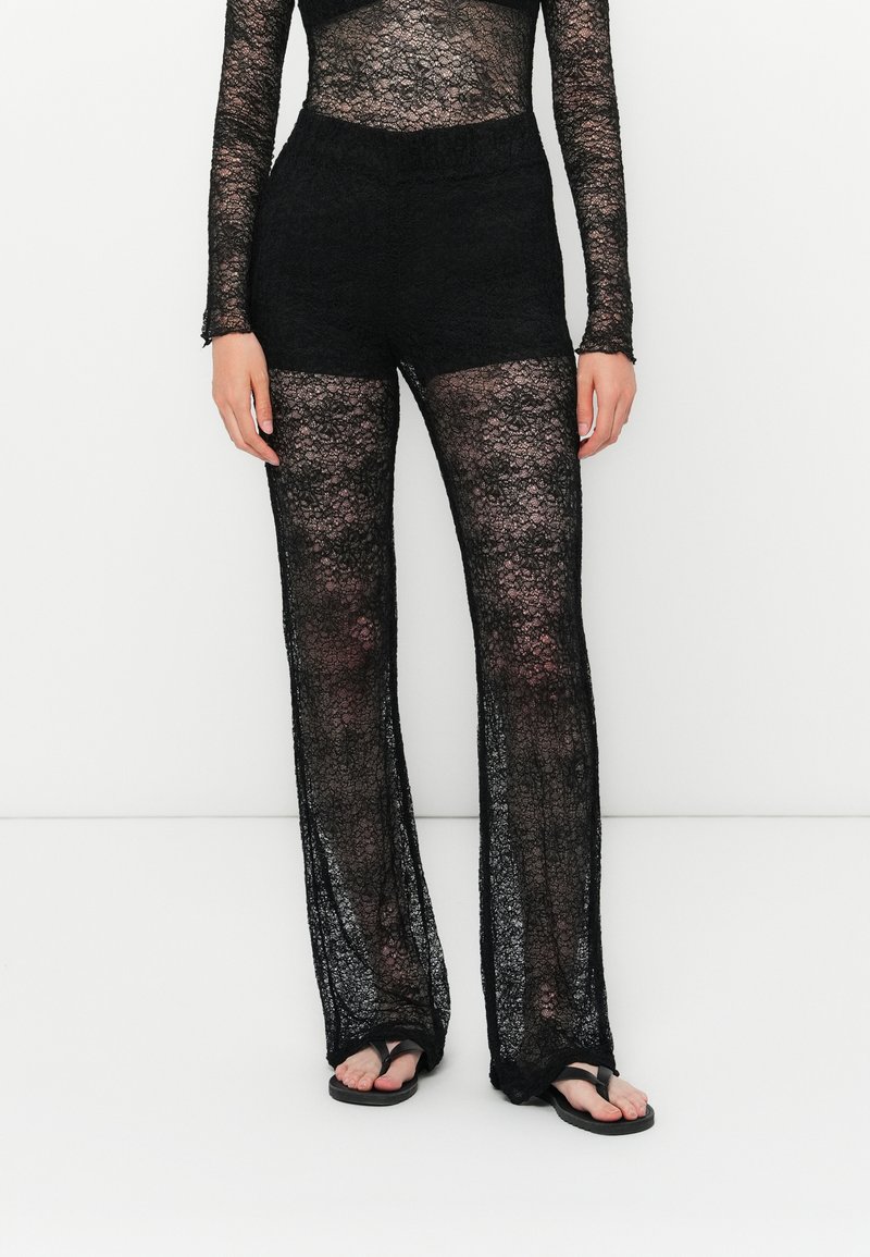 Woman wearing black lace sheer trousers with solid black shorts underneath and black flip-flop sandals, standing against white background.
