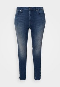 Dark blue denim jeans with a fitted design, five pockets, contrasting stitching, and a zippered hem detail at the ankles.