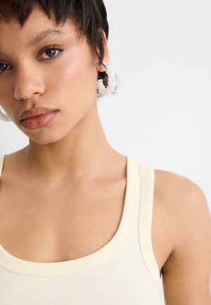 Mladá osoba s krátkými tmavými vlasy nosí velké duhové kruhové náušnice a krémový žebrovaný nátělník na jednoduchém světlém pozadí.