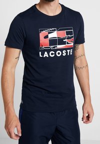Námořnická modrá bavlněná tričko s grafickým designem v červené a bílé barvě a výrazně zobrazeným slovem "LACOSTE".