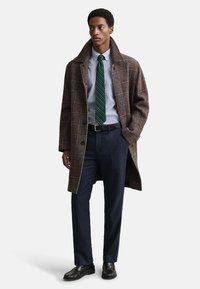 Ruskea ruudullinen ylipaita, jossa on suuri kaulus, päällä vaaleansininen paita ja vihreäraitaista solmiota. Tumma housut ja mustat kengät täydentävät asun.