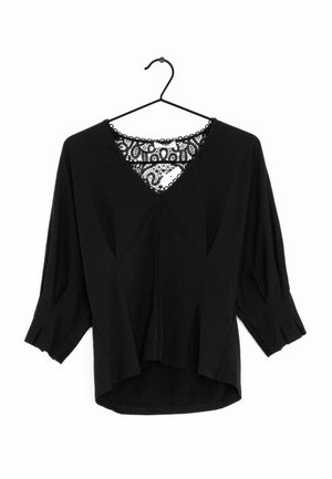Zwarte blouse met lange mouwen, V-hals en kantdetail op de rug, hangend aan een zwarte hanger tegen een witte achtergrond.