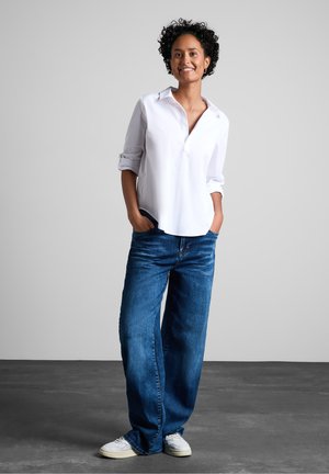 Vit långärmad skjorta med knappar och uppvikta ärmar, i kombination med löst sittande blå denimjeans och vita sneakers, stående mot en neutral bakgrund.
