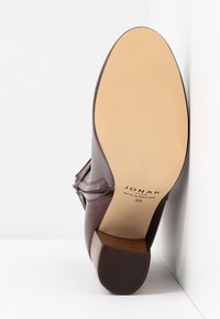 Zapato de cuero marrón con tacón cuadrado, que presenta una superficie suave y acentos decorativos en los costados. La suela es beige con textura de goma. Talla 38.
