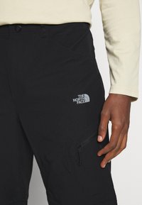 Gros plan sur une personne portant un pantalon noir The North Face avec une poche zippée, une chemise beige à manches longues, et une main droite visible reposant sur le côté.