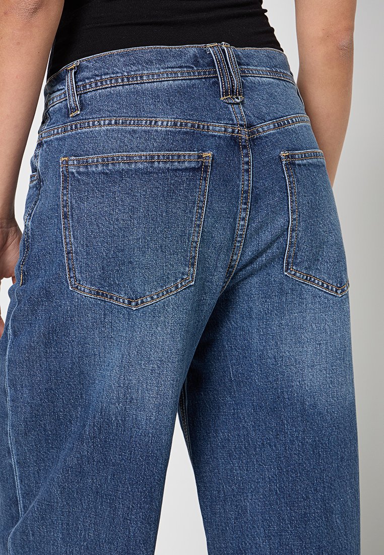 Rückansicht einer Person in blauen Jeans mit Gesäßtaschen und einem schwarzen Oberteil vor einem einfarbigen Hintergrund.