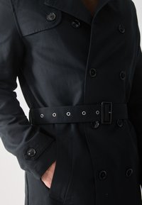 Abrigo trench negro de doble botonadura con cintura ceñida, que presenta botones brillantes, bolsillos laterales y una textura suave.