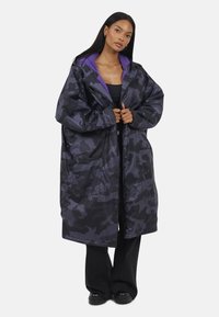 Donna che indossa un ampio cappotto lungo in camuffamento nero e viola con fodera in pile viola, pantaloni neri e scarpe nere, in piedi su uno sfondo bianco.