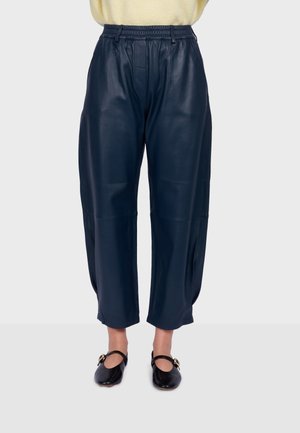 Pantalon en cuir bleu marine ample avec taille élastique porté avec des ballerines Mary Jane noires sur une personne debout sur un fond uni.