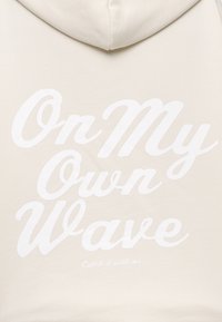 Beige hoodie med en mjuk textur, med stor vit text: "On My Own Wave" och en mindre rad nedan, "Catch it with me."