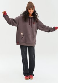 Veľká šedá mikina s červenými detailmi, mäkká tkanina, klokanie vrecko, vyšívaný detail, spojená s tmavými džínsami a červenými teniskami.