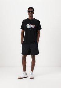 Zwarte katoenen t-shirt met kleurrijk grafisch ontwerp van golven en palmbomen. Gecombineerd met zwarte shorts en witte sneakers.