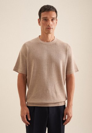 Beige gebreide kortemouw crew neck trui met een rechte pasvorm, ribbels aan de mouwen en zoom, gedragen over marineblauwe broeken. Zachte textuur.