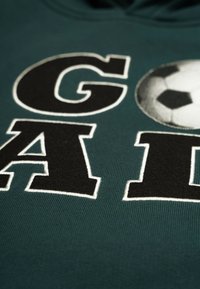 Mörkgrön hoodie med stora svarta bokstäver som står "GOAL" och en präglad fotbollsgrafik, med texturerad tygdetalj synlig.