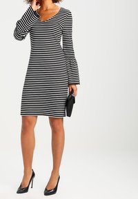 Raidallinen musta-valkoinen mekko V-mallisella kauluksella, pitkähihaisilla kellotetuille hihoille ja sopivalla siluetilla. Yhdistetty mustien korkokenkien ja clutch-laukun kanssa.